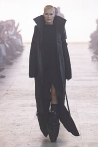 Model walks a runway in an all-black, oversized coat with a high collar and knee-high boots.