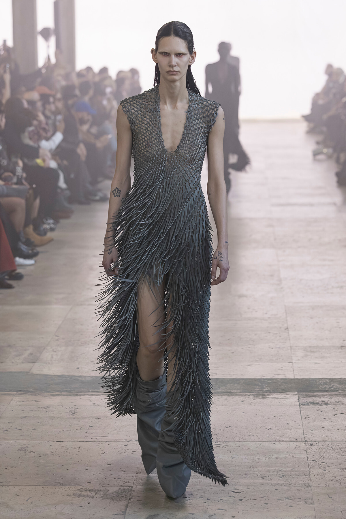 Model walks a fashion runway in a dark, fringed gown with a mesh-like bodice; audience blurred in the background.