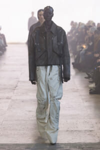 Model walks the runway in a dark fur jacket with front zips and harness straps, paired with light gray cargo pants.