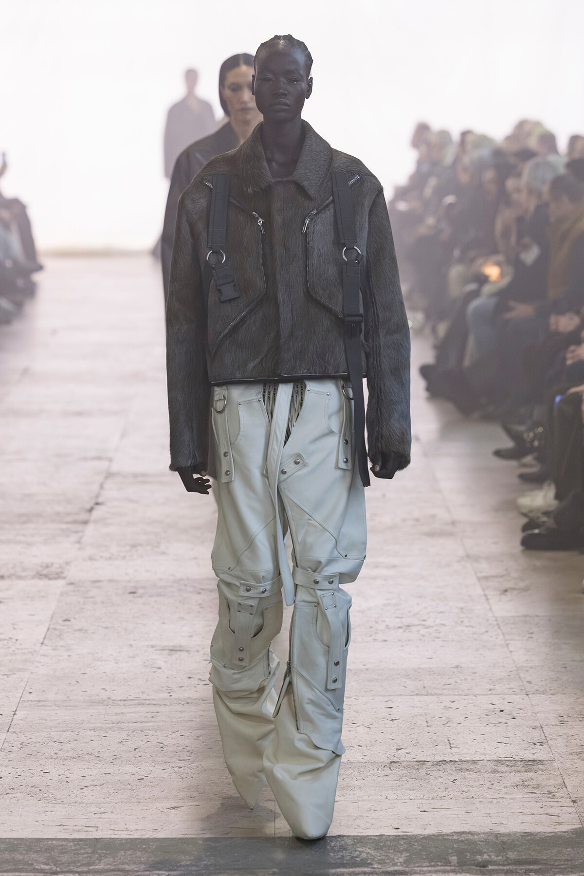 Model walks the runway in a dark fur jacket with front zips and harness straps, paired with light gray cargo pants.