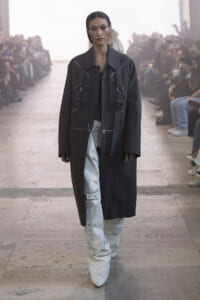 Model walks a runway in a long black coat with white cargo-style pants and chunky white boots, audience on both sides.