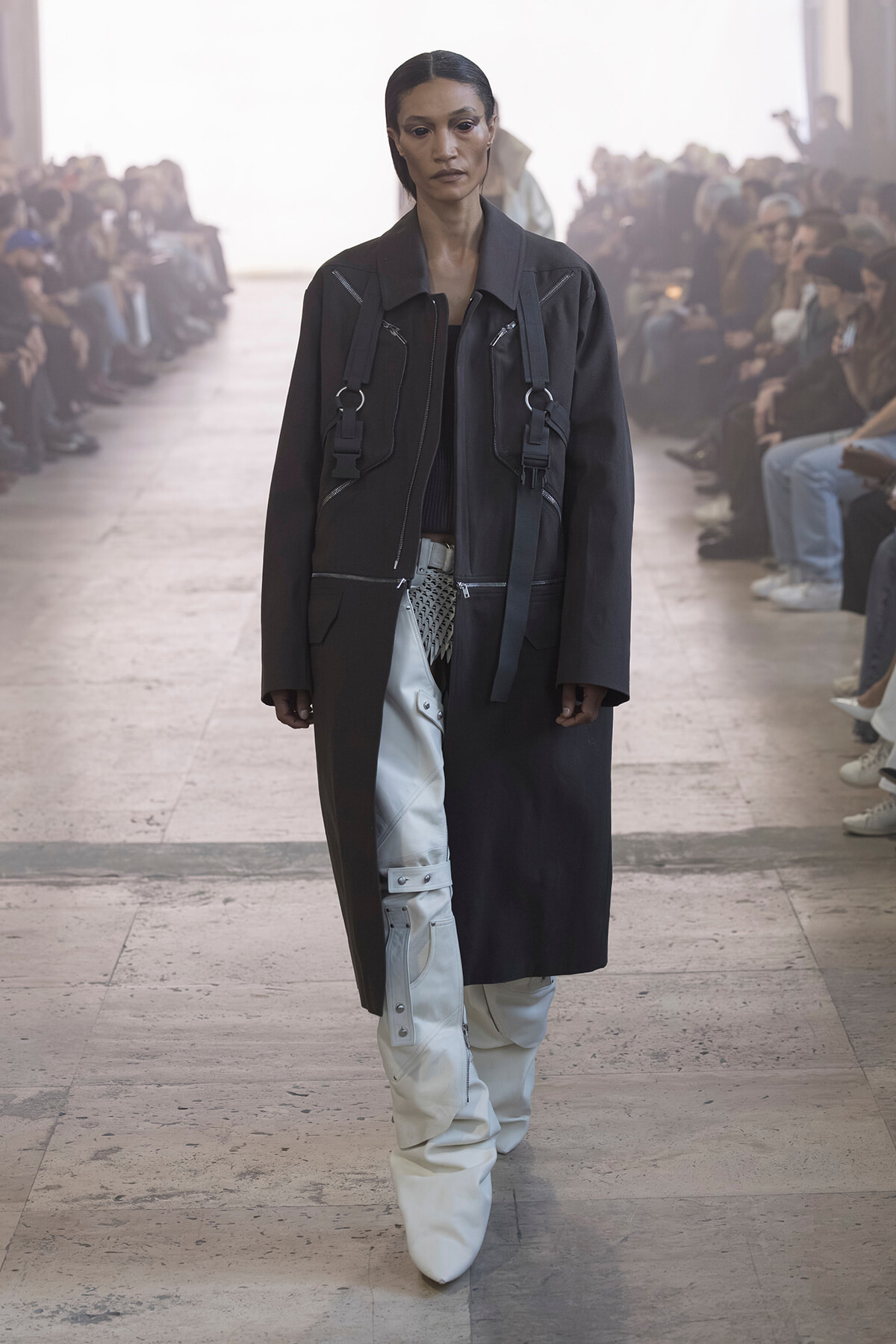 Model walks a runway in a long black coat with white cargo-style pants and chunky white boots, audience on both sides.