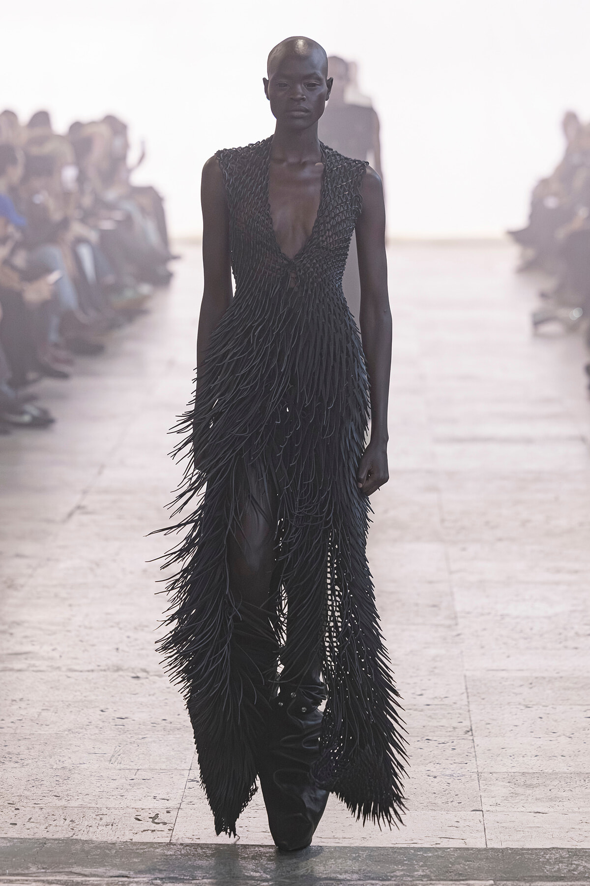 Model walking runway in a long black fringe dress with a plunging neckline, feather-like strands cascading down the gown, audience on both sides.