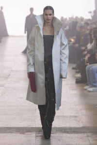 Model walks the runway in a pale oversized coat over a black top, holding a burgundy clutch as spectators watch.