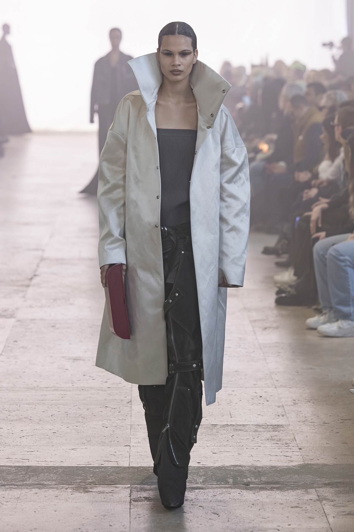Model walks the runway in a pale oversized coat over a black top, holding a burgundy clutch as spectators watch.