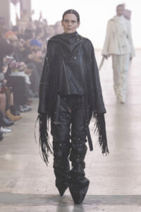 Model on a fashion runway wearing a black fringe leather jacket and pants with metal zippers; audience in background on both sides.