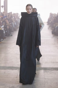 Model walks the runway in a long black coat with an oversized, high collar; audience visible in the background.