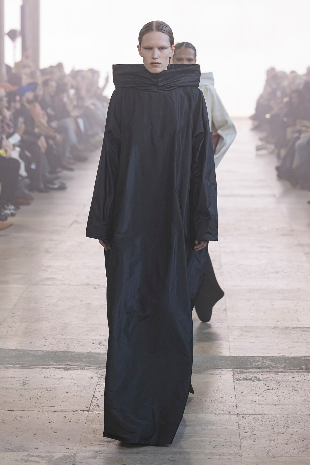 Model walks the runway in a long black coat with an oversized, high collar; audience visible in the background.