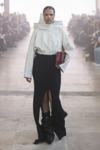 Model walk on a runway in a white oversized top with a high collar, black slit-knit skirt, and black slouch boots, holding a red clutch.