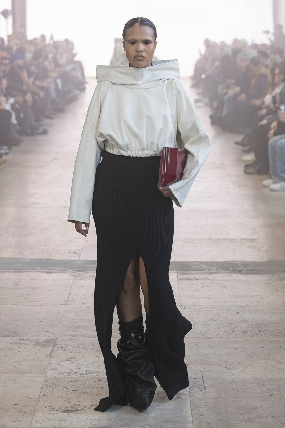 Model walk on a runway in a white oversized top with a high collar, black slit-knit skirt, and black slouch boots, holding a red clutch.