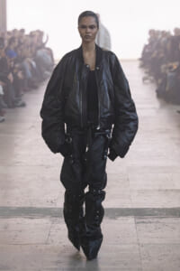 Model on a runway wearing an oversized black leather bomber jacket and matching baggy pants with metal hardware.