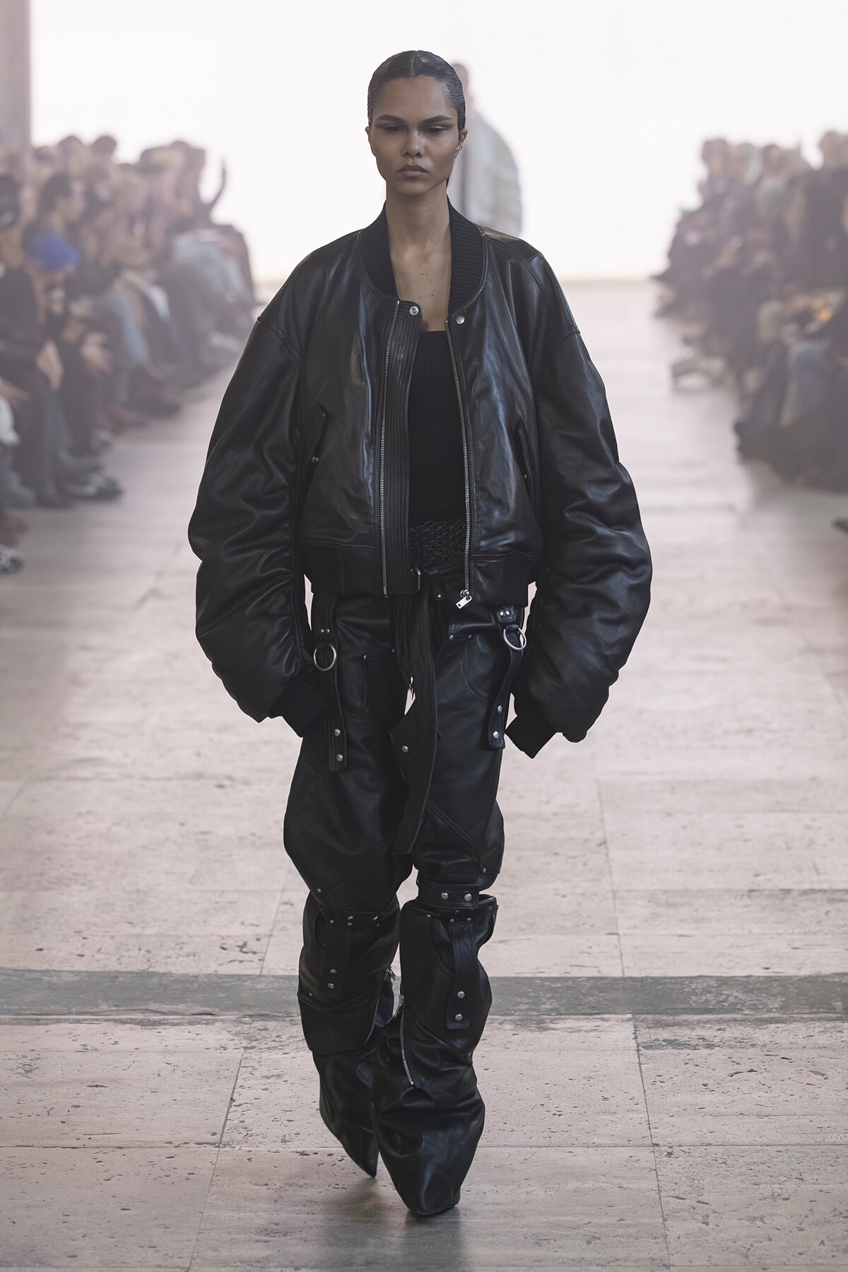 Model on a runway wearing an oversized black leather bomber jacket and matching baggy pants with metal hardware.