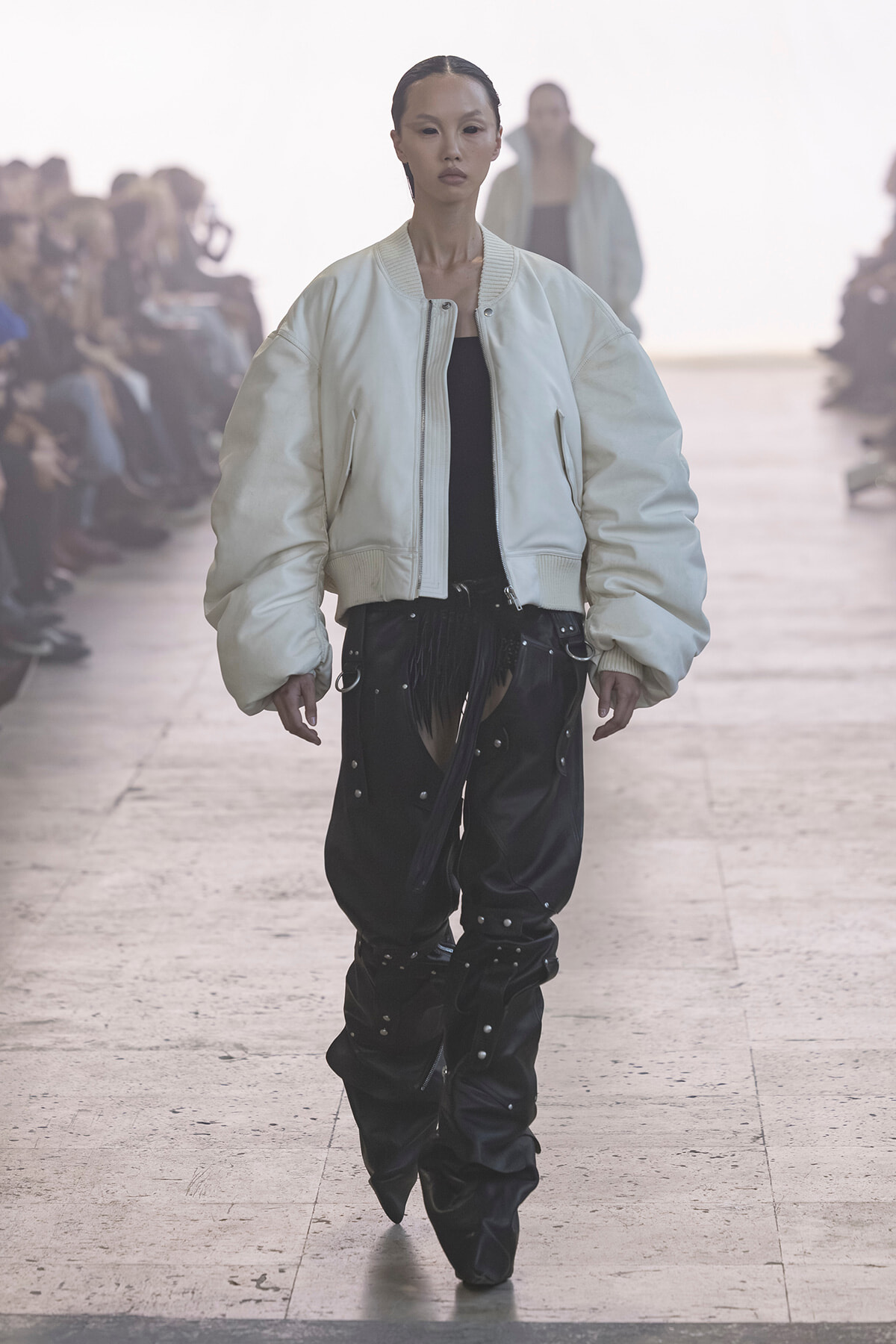 Model walking the runway in an oversized white bomber jacket and black studded pants, audience blurred in the background on a fashion show runway.