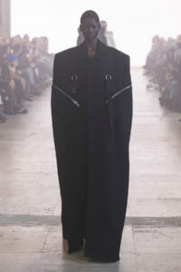 Model walking down a runway in an oversized black coat with metallic zippers and strap details.