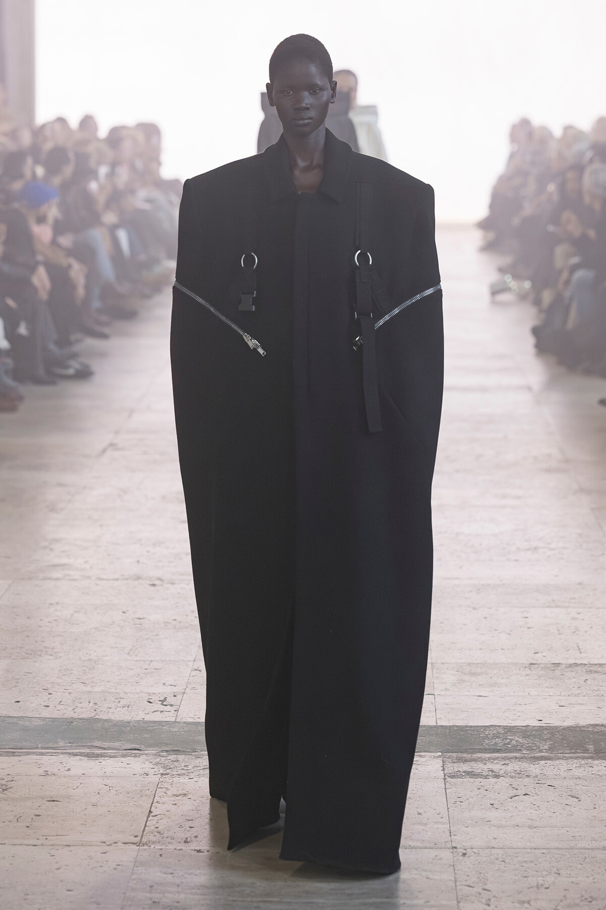 Model walking down a runway in an oversized black coat with metallic zippers and strap details.