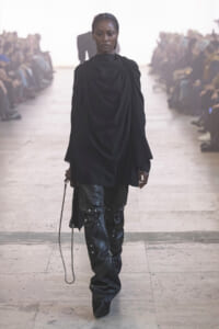 Model walks the runway in a black draped top and studded leather pants, with a blurred audience on both sides.