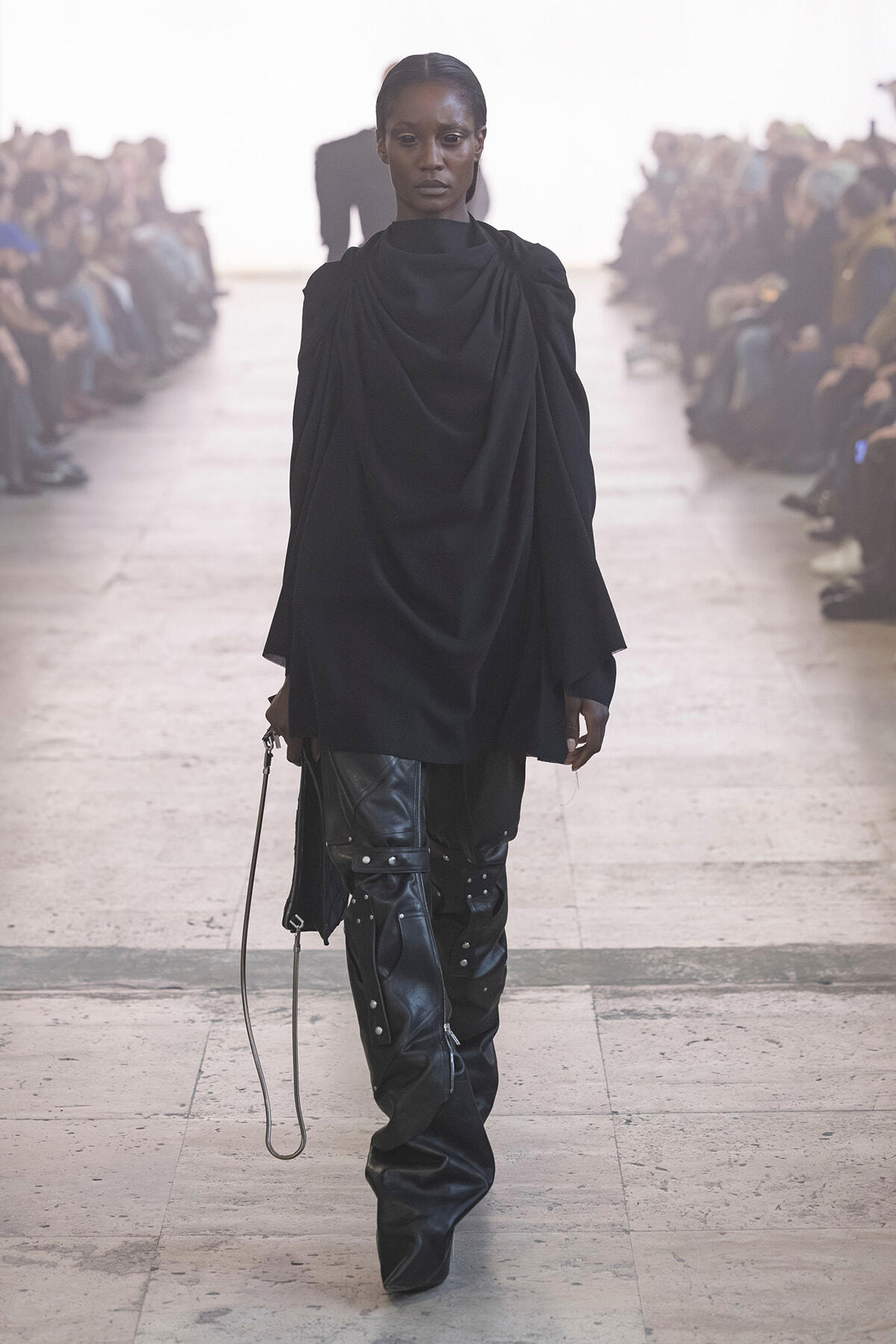 Model walks the runway in a black draped top and studded leather pants, with a blurred audience on both sides.