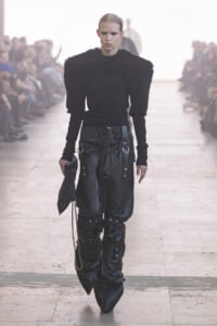 Model walking a runway in an all-black outfit: fuzzy-textured shoulder top and oversized leather pants with hardware, clutch in hand