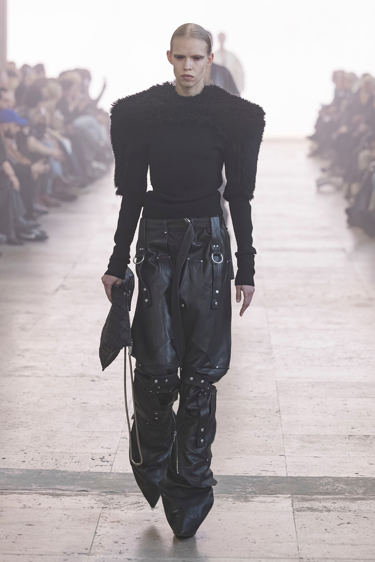 Model walking a runway in an all-black outfit: fuzzy-textured shoulder top and oversized leather pants with hardware, clutch in hand