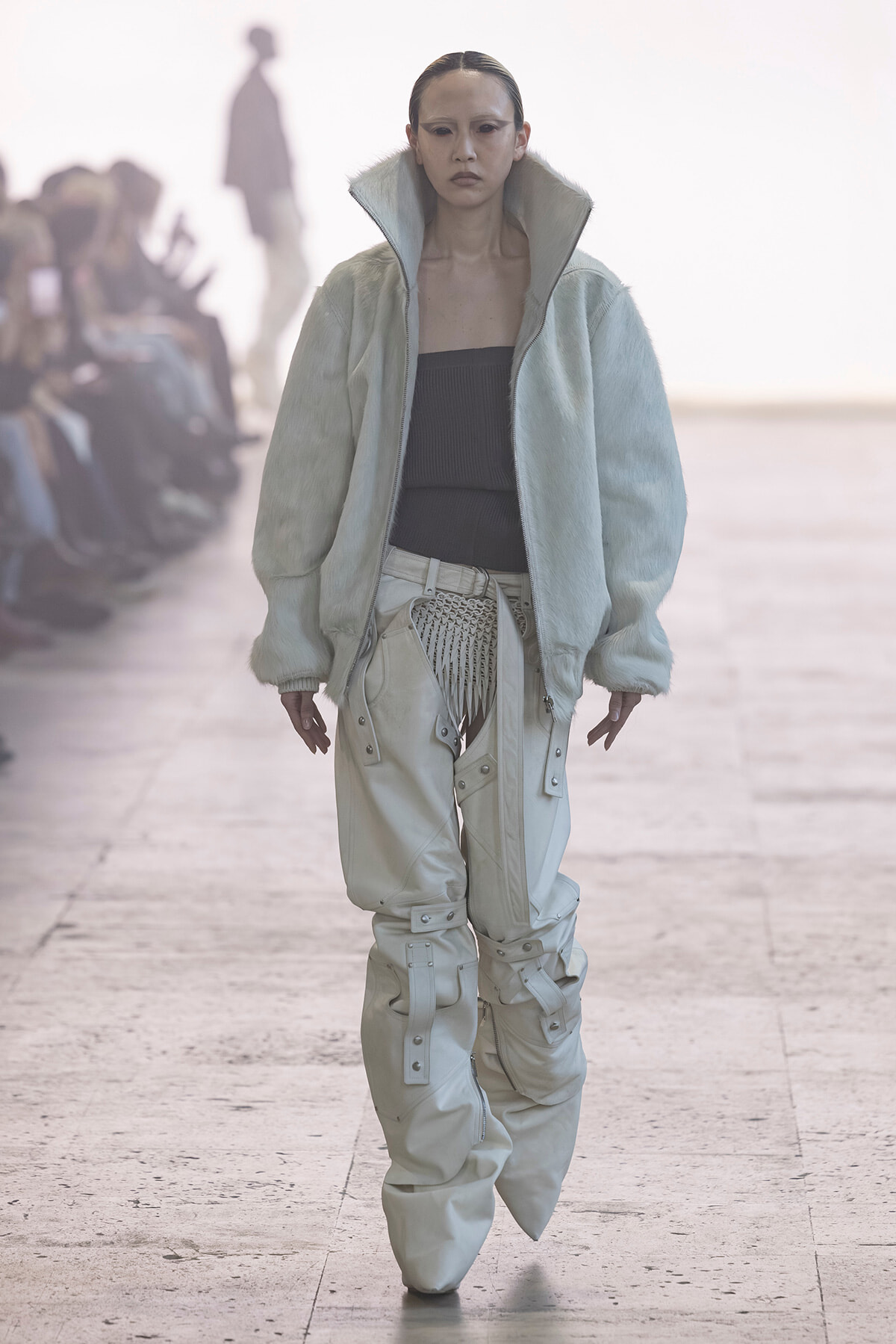 Female runway model wearing an oversized pale mint fur-like jacket over a black tube top and cream cargo pants with buckles and straps on a catwalk