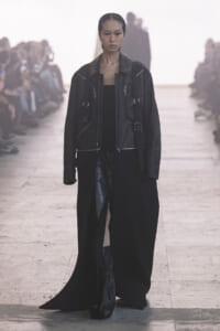 Model walking the runway in a black leather jacket and long black coat, audience blurred in the background.