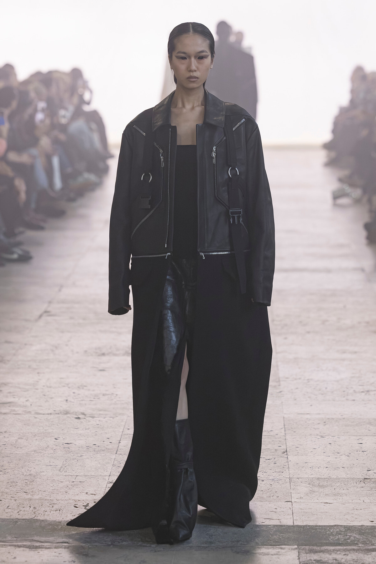 Model walking the runway in a black leather jacket and long black coat, audience blurred in the background.