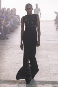 Model walks a runway in a black sleeveless dress with cutouts; audience blurred in the background.
