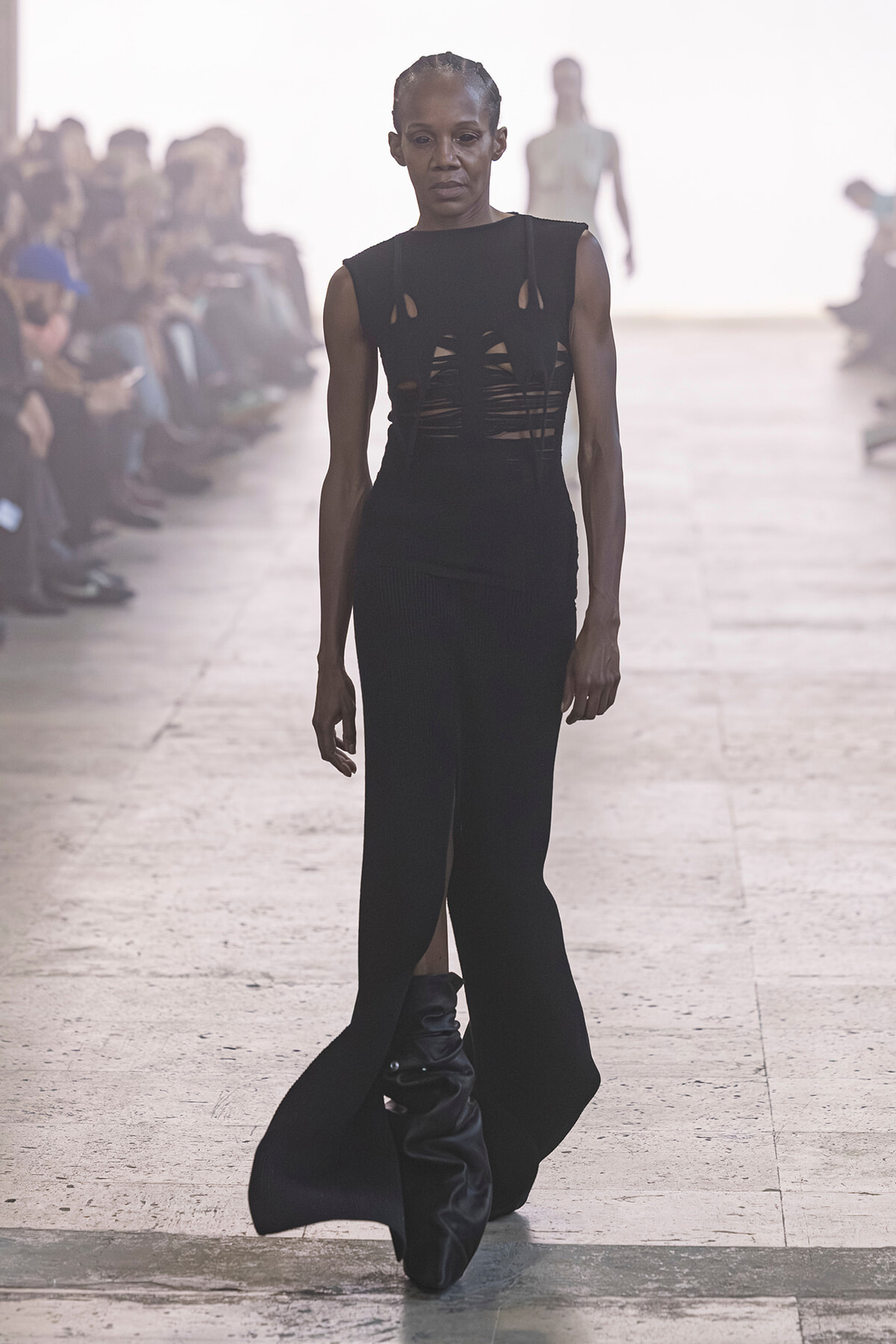 Model walks a runway in a black sleeveless dress with cutouts; audience blurred in the background.