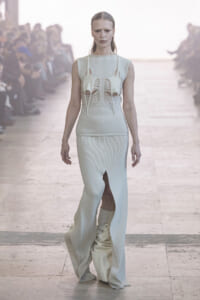 Model walks runway in a pale ivory, sculptural knit gown with cutouts at the chest and long, flared skirt; audience blurred in background.