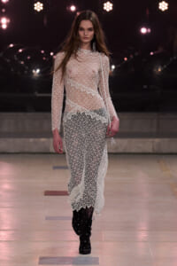 Model walks a runway in a sheer white lace dress over dark pants, paired with black studded knee-high boots, illuminated by stage lights in the background.