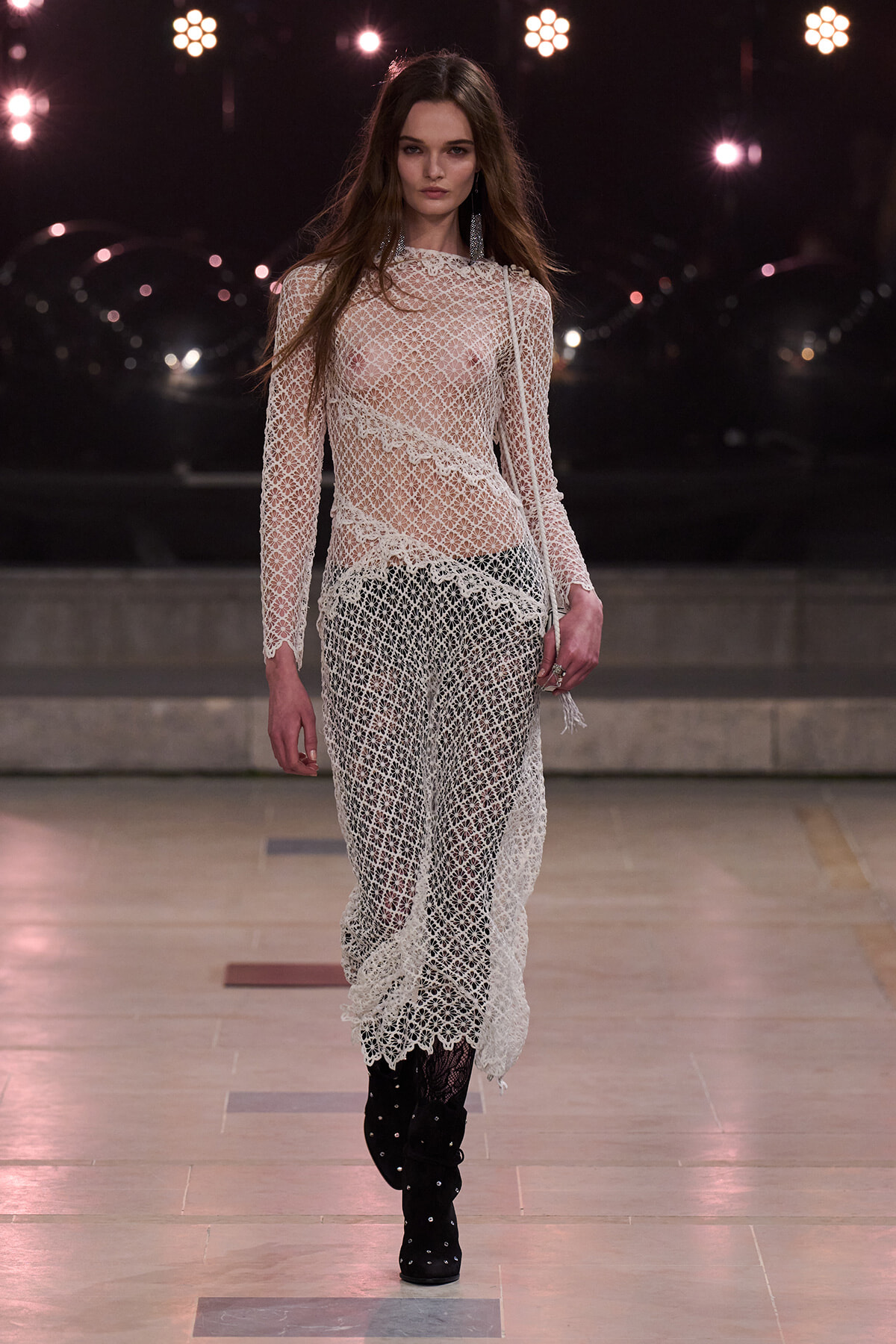 Model walks a runway in a sheer white lace dress over dark pants, paired with black studded knee-high boots, illuminated by stage lights in the background.