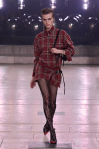 Model struts down a runway in a red plaid shirts-dress with a black belt, fishnet floral tights, and red-heeled pumps while carrying a black bag.
