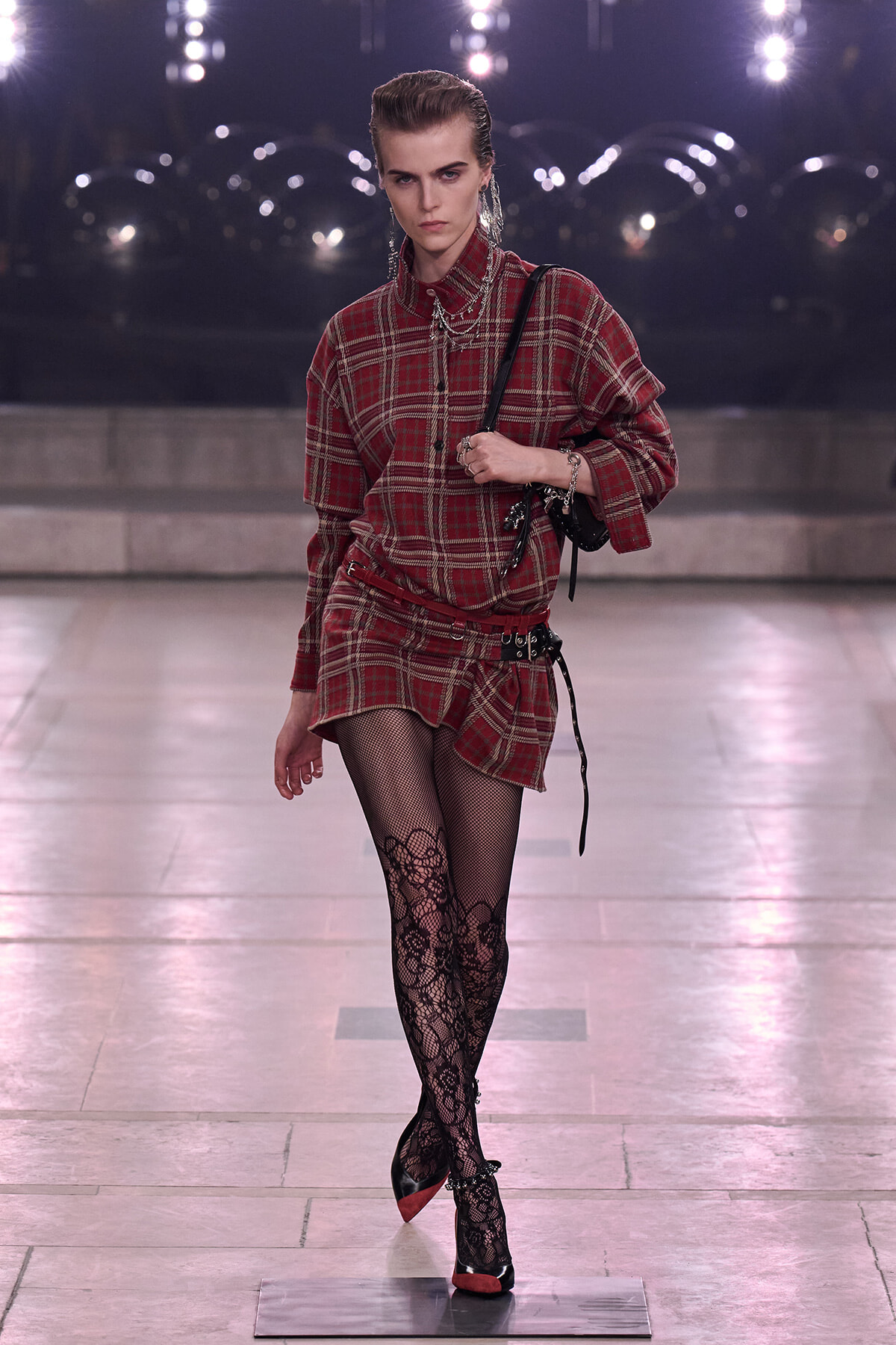 Model struts down a runway in a red plaid shirts-dress with a black belt, fishnet floral tights, and red-heeled pumps while carrying a black bag.