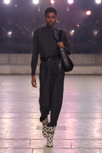 Model walks a fashion runway in a dark pinstripe suit with a tie, black belt, and a large black handbag, paired with white polka-dot boots.