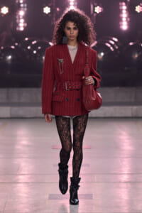 Runway model in a burgundy pinstripe blazer with belt, black lace tights, and black ankle boots, carrying a red handbag under bright stage lights.