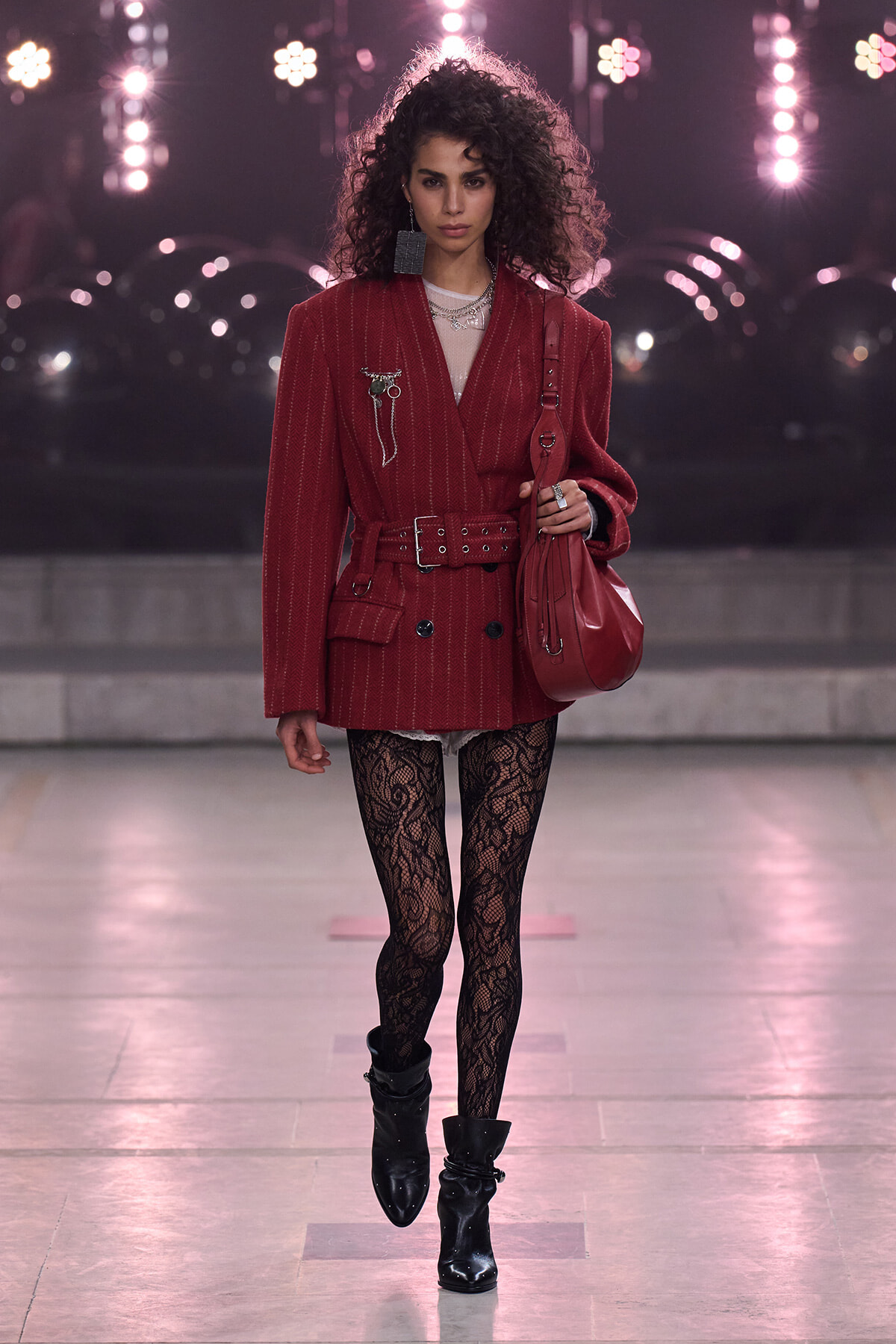 Runway model in a burgundy pinstripe blazer with belt, black lace tights, and black ankle boots, carrying a red handbag under bright stage lights.
