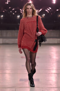 Model walking a runway in an oversized red knit sweater dress with a white pattern, paired with black fishnet tights and studded black ankle boots, carrying a black shoulder bag. Under pink runway lights.