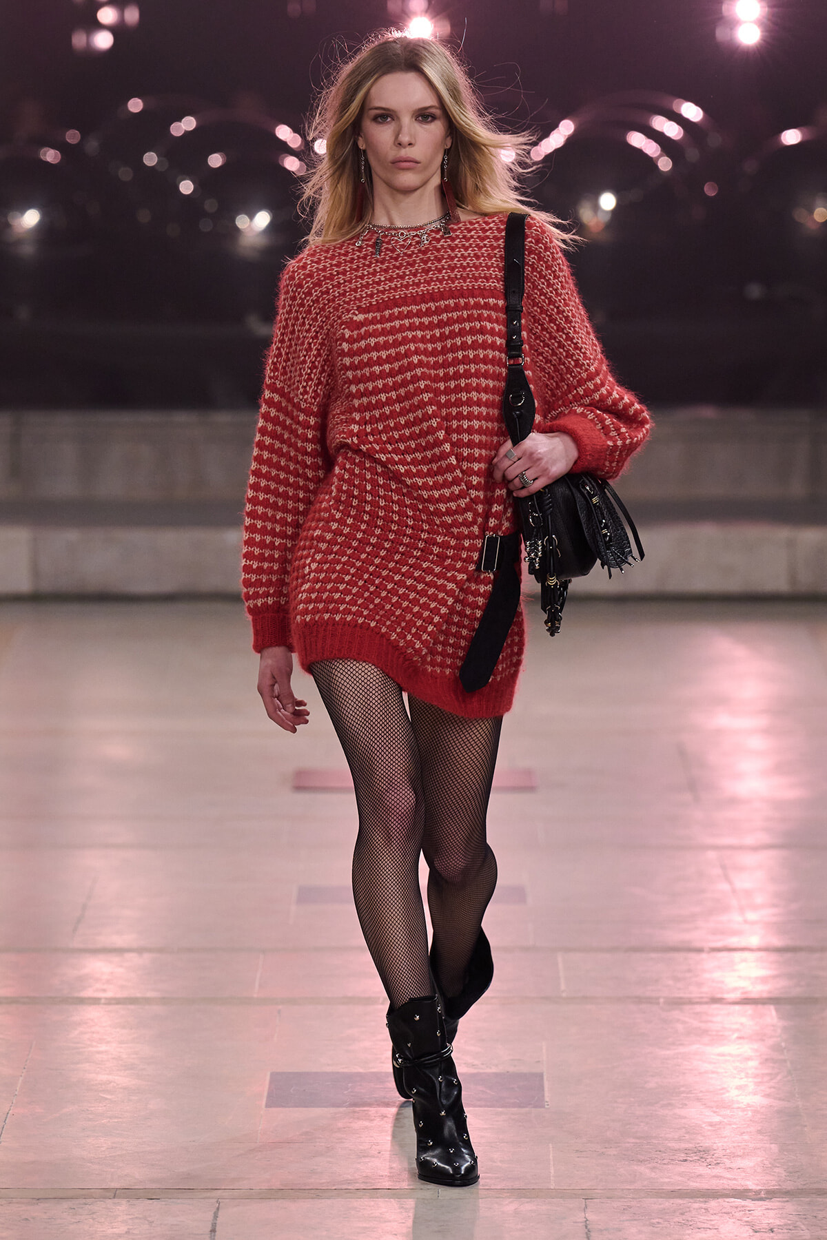 Model walking a runway in an oversized red knit sweater dress with a white pattern, paired with black fishnet tights and studded black ankle boots, carrying a black shoulder bag. Under pink runway lights.