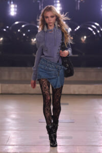 Model walking a runway at night in a lavender oversized hoodie, layered denim wrap skirt, black lace tights, and studded heeled boots while carrying a black bag.