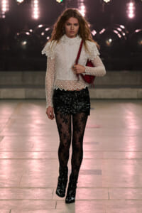 Model walking a runway in a white lace high-neck top with scalloped edges, a black studded leather mini skirt, lace tights, black heeled ankle boots, and a red shoulder bag.