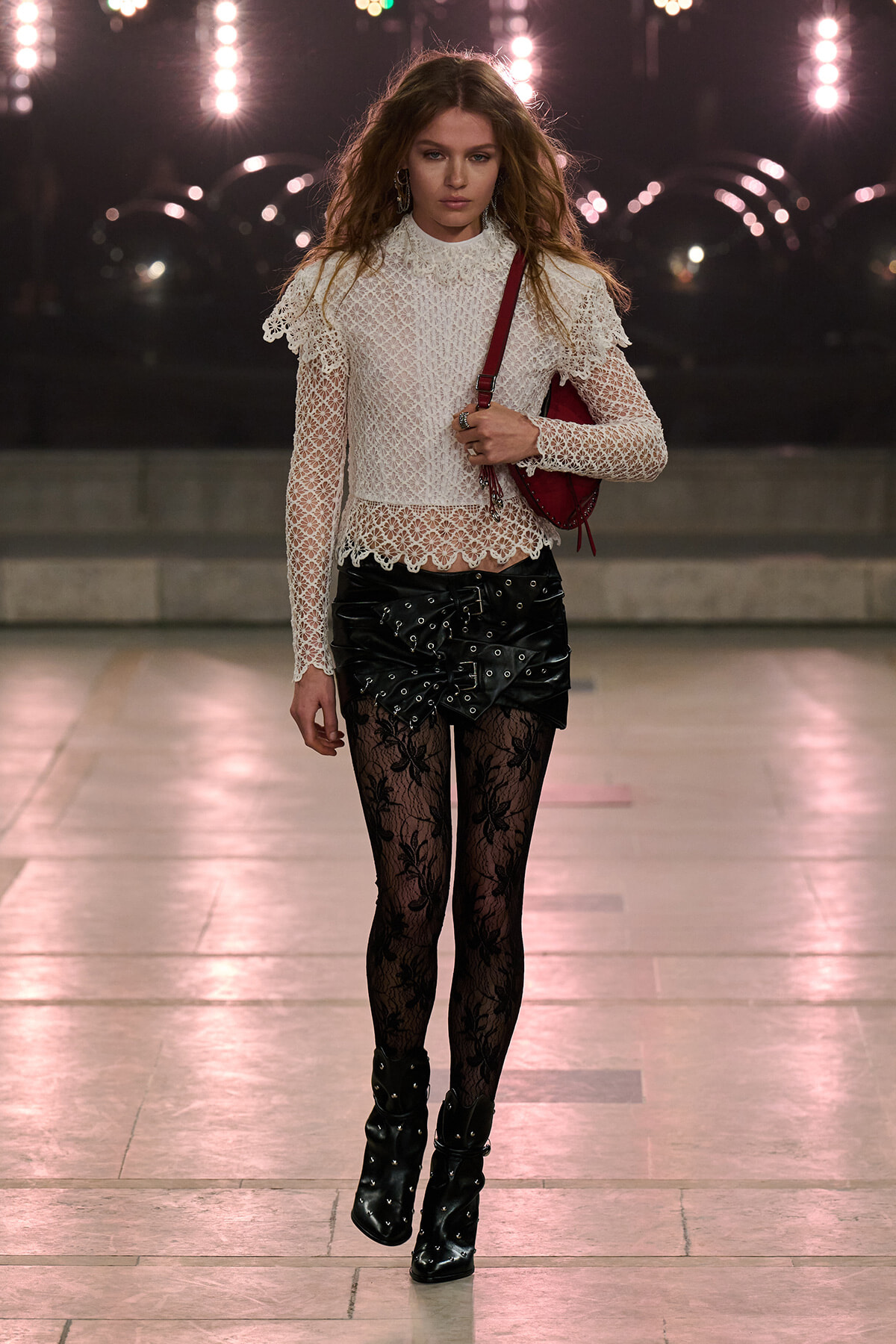 Model walking a runway in a white lace high-neck top with scalloped edges, a black studded leather mini skirt, lace tights, black heeled ankle boots, and a red shoulder bag.