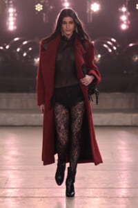 Model walking a runway in a burgundy coat over a sheer black top, lace tights, and black ankle boots during a fashion show.