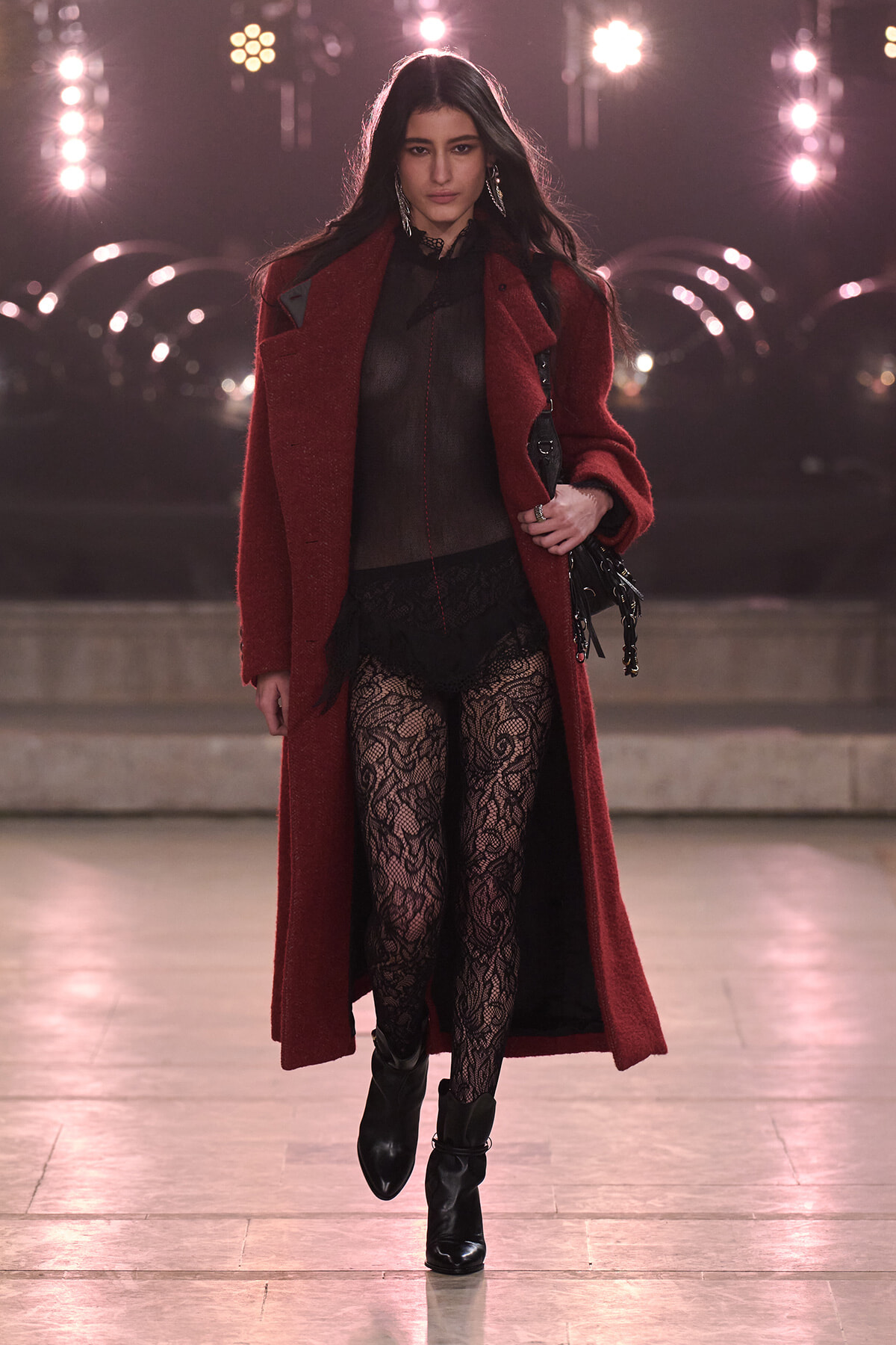 Model walking a runway in a burgundy coat over a sheer black top, lace tights, and black ankle boots during a fashion show.
