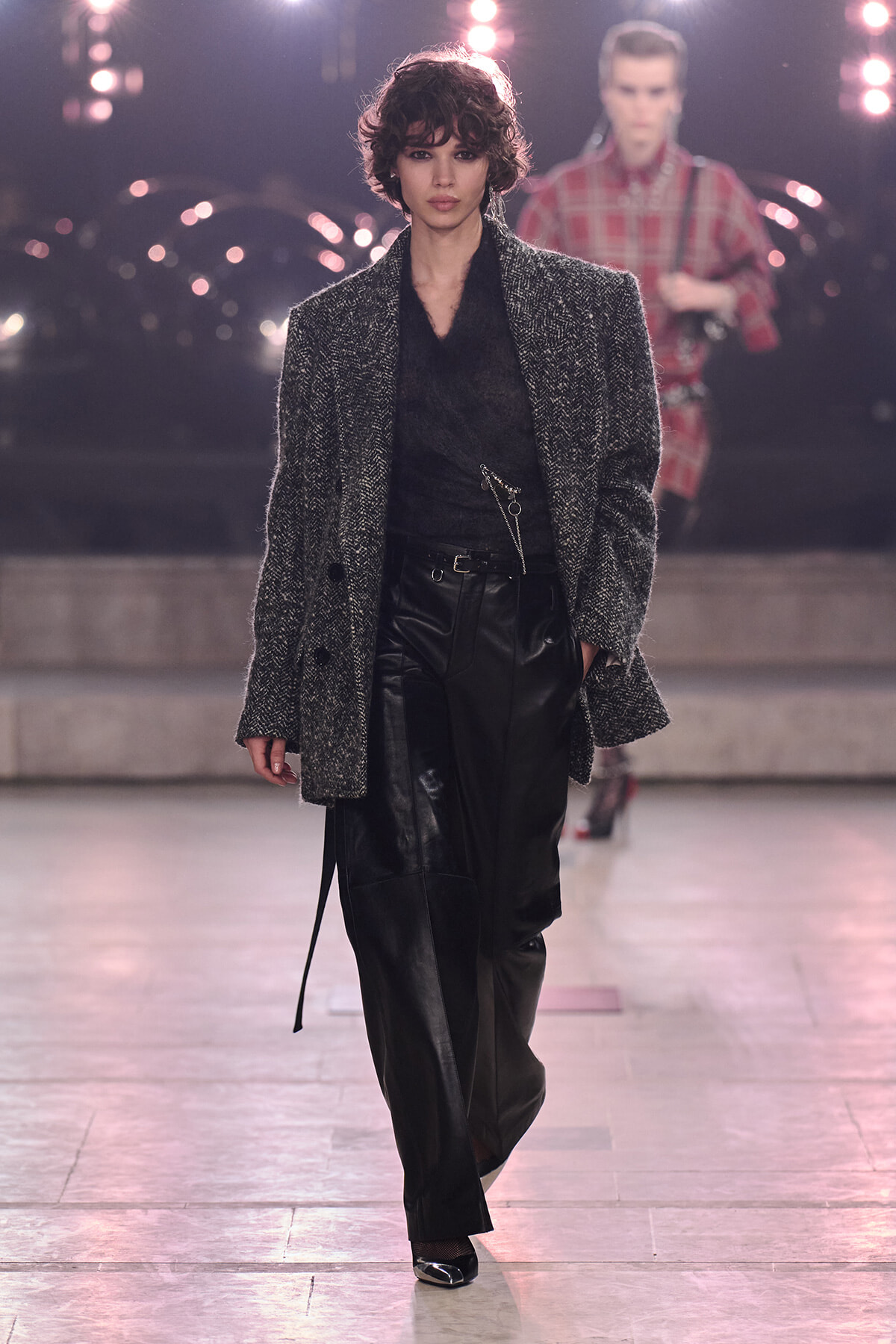 Model walks a runway in a textured gray overcoat over a sheer black top and shiny black leather pants, under bright stage lights.