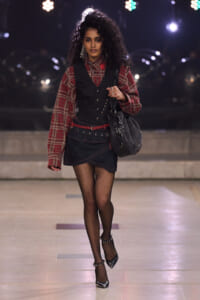Model walking a fashion runway at night in a red plaid blouse, black vest, and denim skirt with fishnet tights and black heels carrying a black bag.