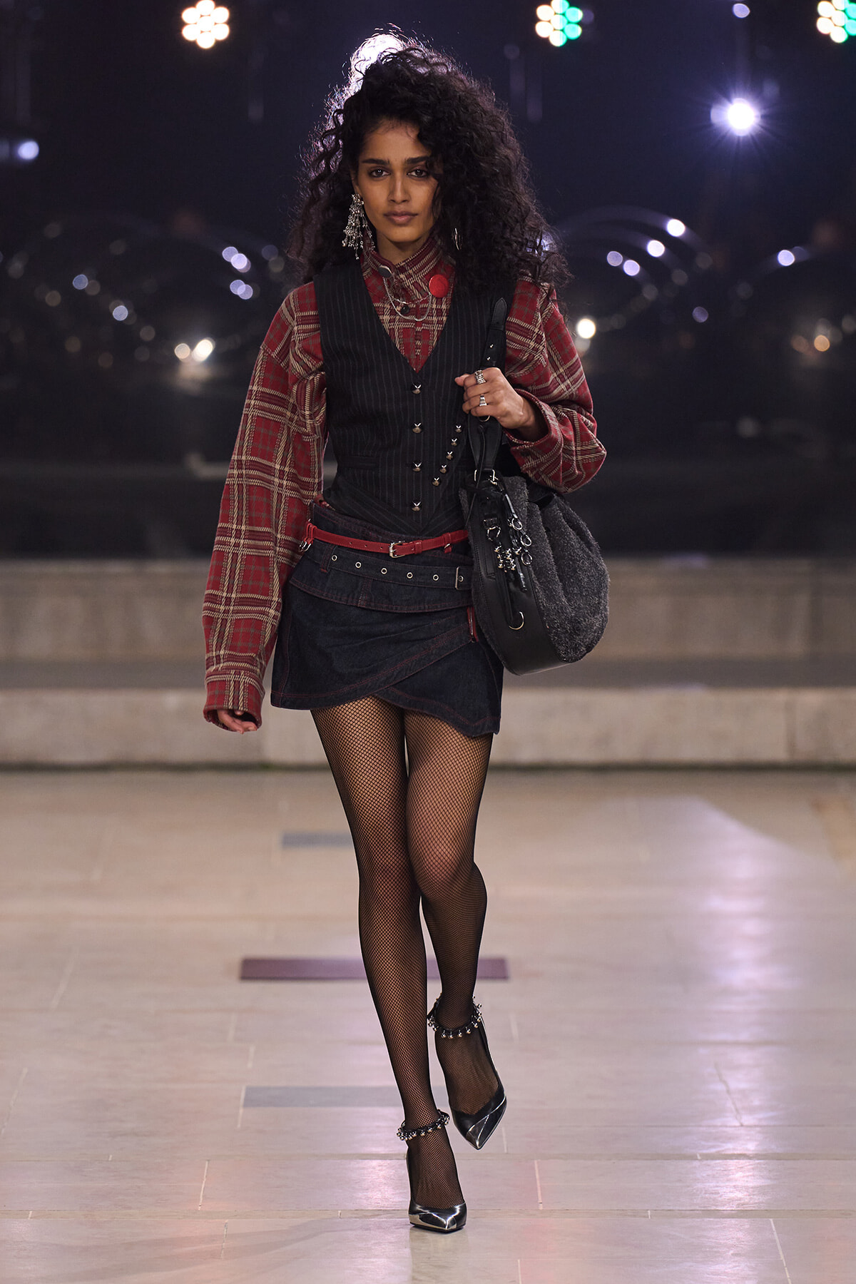 Model walking a fashion runway at night in a red plaid blouse, black vest, and denim skirt with fishnet tights and black heels carrying a black bag.