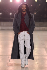Runway model walking in a long gray overcoat over a burgundy knit, light gray pants, and white boots.