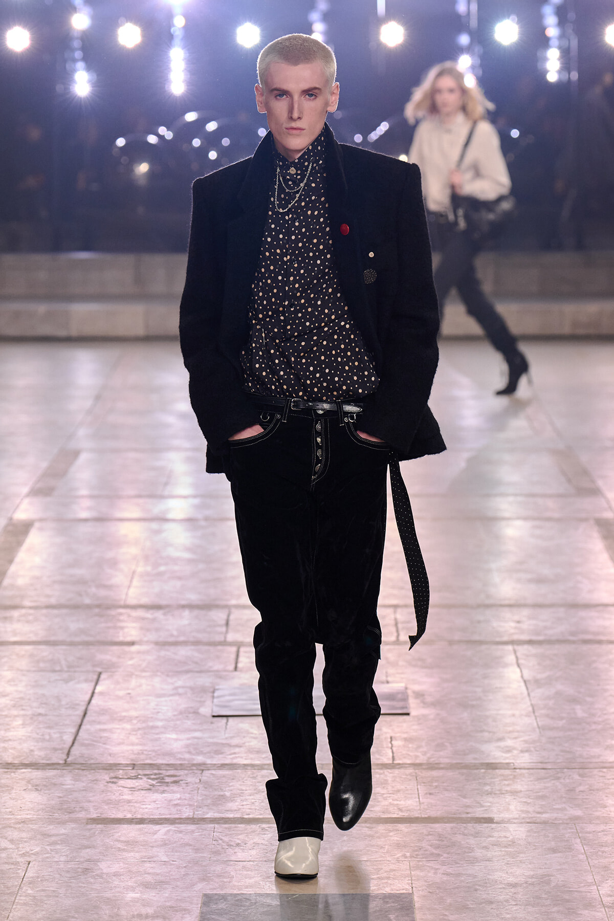 Male model struts a fashion runway in a black textured blazer, polka-dot shirt, layered chains, and black velvet pants with two-tone boots.