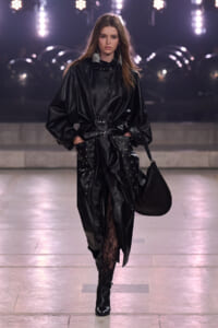 Model strides down a runway in a shiny black leather coat with a belt, oversized sleeves, riveted pockets, and a large black handbag.