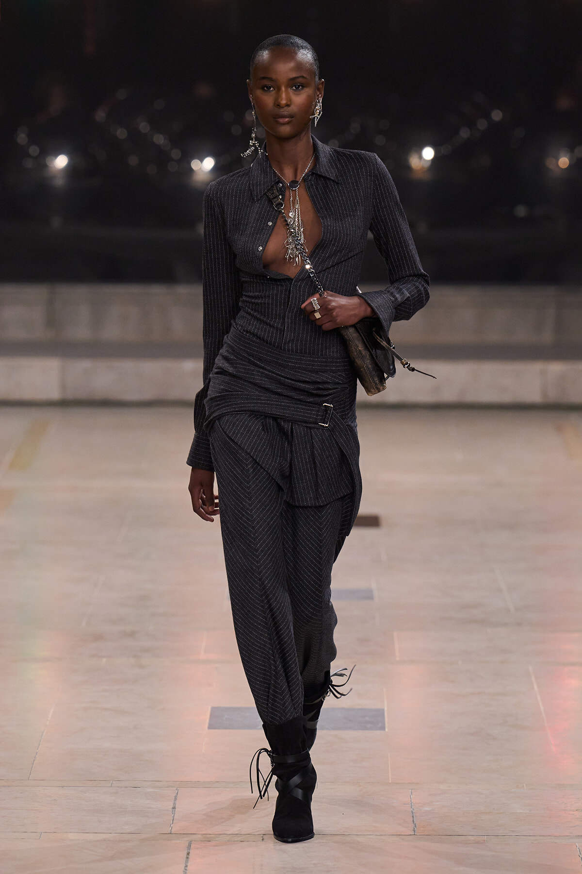 Model walking on a runway in a dark pinstripe outfit with an open chest shirt, layered silver jewelry, and a small bag slung over the shoulder; knee-high lace-up boots complete the look.